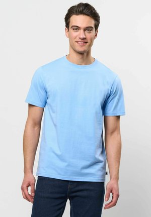 Jonge man met bruin haar die een lichtblauw T-shirt met korte mouwen en donkere spijkerbroek draagt, staand tegen een effen lichte achtergrond.