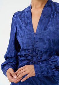 V-neck blue satin blouse with textured pattern, puff sleeves, gathered front, and buttoned cuffs. Smooth finish and elegant drape.
