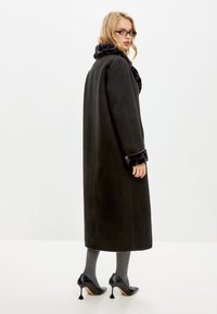 Lange zwarte jas met een pluche zwarte kraag en manchetten, met zijzakken, gedragen met grijze gestructureerde sokken en zwarte hoge hakken.