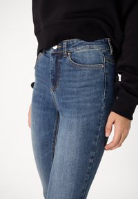 Blå denimjeans med en figursydd design, med framfickor, silverdetaljer och orange sömnad. Matchas med en svart sweatshirt.