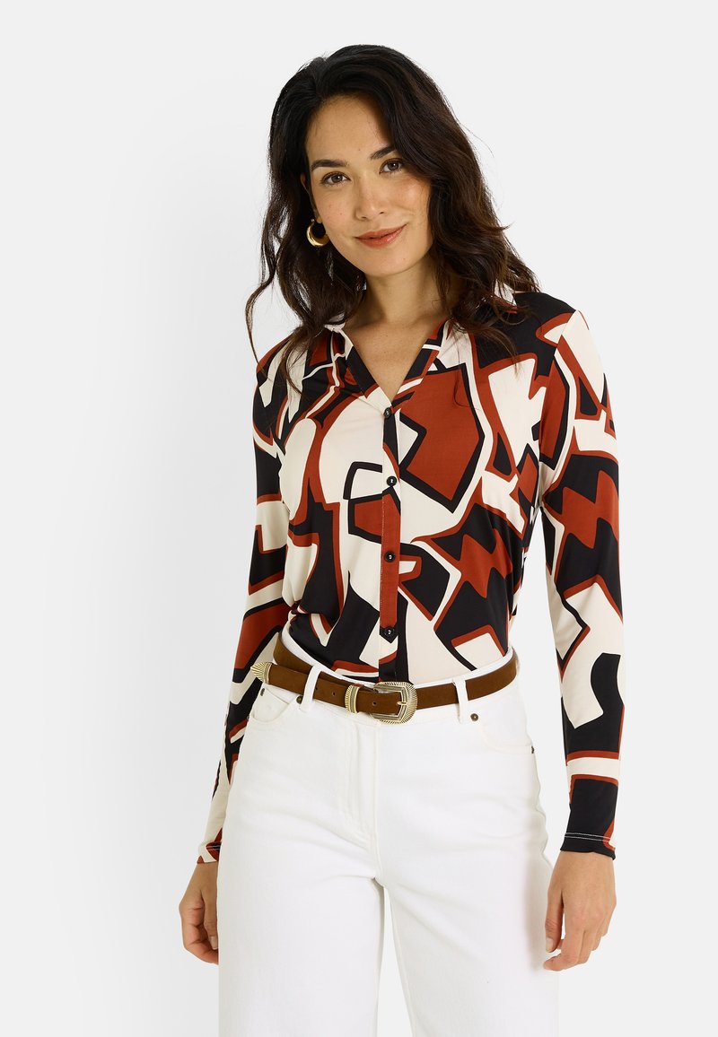 Camisa de manga larga con un atrevido patrón abstracto en rojo, negro y crema. Presenta un cierre de botones en la parte frontal y botones negros. Se lleva con jeans de color claro.