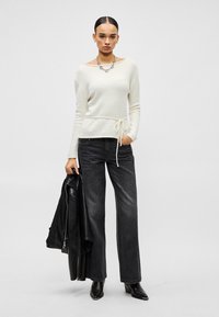 Femme portant un pull blanc à manches longues avec ceinture à la taille, jean noir à jambes larges, bottes noires, tenant une veste en cuir noire dans une main.