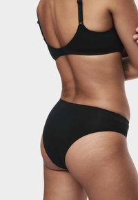 Donna che indossa un reggiseno e slip neri, vista di spalle, con un tatuaggio sul braccio sinistro su uno sfondo neutro.