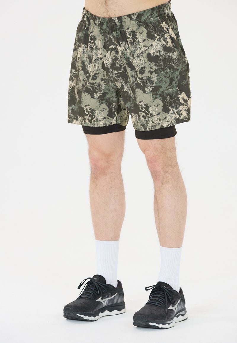 Camouflaged sportieve shorts in donker groen en beige met een elastische tailleband, gecombineerd met zwarte compressieshorts, witte sokken en zwarte sneakers.