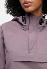 Ljuslila anorakjacka med hög krage, justerbara dragskor och en framficka med horisontell klaff. Mjukt, strukturerat tyg.