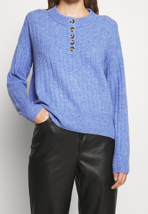 Vrouw draagt een geribbelde lichtblauwe trui met vijf knopen bij de halslijn en zwarte leren broek, hand in zak.
