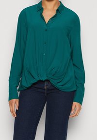 Camisetta lunga teal con fronte annodato, colletto e design con bottoni. Realizzata in un tessuto morbido con una vestibilità comoda. Abbinata a jeans scuri.
