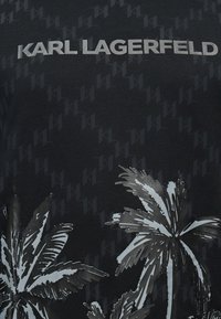 Μαύρο πουκάμισο με επαναλαμβανόμενο διακριτικό μοτίβο "KL", μεγάλο ασημένιο κείμενο "KARL LAGERFELD" στην κορυφή και δύο ασημένια γραφικά φοίνικες στο κάτω μέρος.