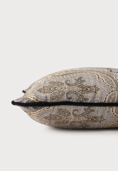 Coussin décoratif avec motif cachemire beige et brun complexe et passepoil noir sur le bord, sur un fond uni clair.