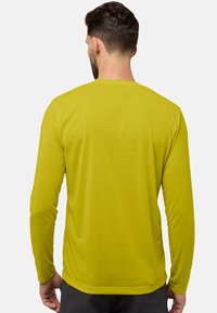 Man met kort donker haar en een baard, gekleed in een eenvoudig gele longsleeve shirt, met zijn rug naar de camera gekeerd tegen een effen lichte achtergrond.
