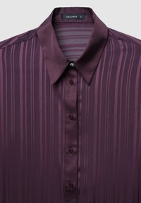 Lila gestreifte Bluse mit Satin-Kragen und Knopfverschluss, versehen mit einem kleinen dekorativen Juwel in der Mitte des Kragens.