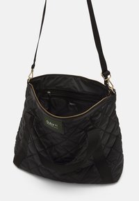 Sac cabas noir matelassé avec un extérieur texturé, une fermeture éclair dorée et deux anses noires ; comprend des compartiments intérieurs et un patch logo vert.