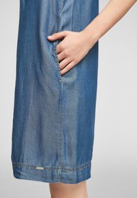 Denimové šaty s boční kapsou, v světlé modré barvě, jemné textuře a kontrastním prošitím podél švů.