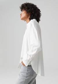 Weißes Langarmbluse mit hohem Kragen, gefälteltem Schulterdetail und geknöpften Manschetten. Kombiniert mit grauen Jeans, von der Seite gezeigt.