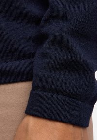 Dunkelblaues Strickpulloverbündchen mit strukturiertem Rippenmuster, glatter Oberfläche und tailliertem Design. Hand am Rand sichtbar.