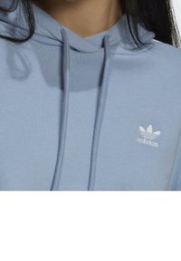 Ljusblå hoodie i mjukt tyg, med en vit Adidas-logotyp på vänster bröstområde och snörningsdetaljer vid huvan.