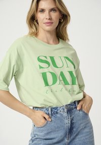Šviesiai žalia trumpomis rankovėmis marškinėliai su užrašu "SUN DAY" tamsiai žalia spalva ir "Chill Out" mažesniu šriftu. Suderinti su aukštu juosmeniu džinsiniais šortais.