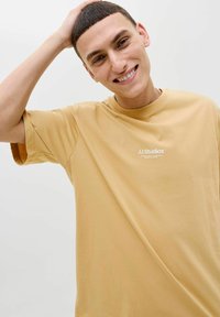 T-shirt à manches courtes, couleur brun clair, en tissu doux, avec le texte "JJ Studios" en blanc, coupe décontractée et encolure ronde.