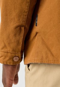 Gros plan sur le bras d'une personne portant une veste marron texturée avec un bouton et une petite étiquette indiquant "EST. 1993", pantalon beige visible.
