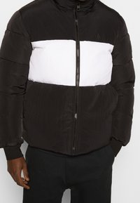 Hombre con chaqueta acolchada negra con una franja blanca horizontal en el pecho, pantalones negros y manos parcialmente en los bolsillos.