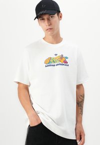 T-shirt bianca in cotone con una grafica di frutta colorata e il testo "ADIDAS ORIGINALS". Abbinata a un berretto nero e jeans scuri.