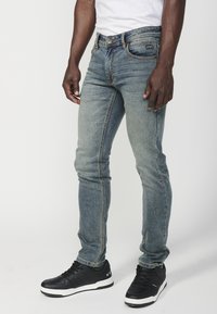 Világoskék, slim fit farmernadrág, kopott textúrával, öt zsebes dizájnnal és kontrasztvarrással. Fekete sportcipővel párosítva.