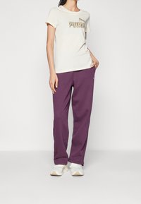 Kvinna iklädd en krämfärgad kortärmad Puma t-shirt, lila träningsoverallsbyxor och vita sneakers, som står med ena handen i fickan mot en vit bakgrund.