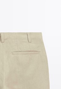 Pantaloni beige in lino con una forma dritta, dotati di due passanti per la cintura, una tasca posteriore e cuciture visibili lungo la vita e i bordi delle tasche.