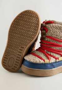 Braune und beige Knöchelstiefel mit roten Schnürsenkeln, einem Obermaterial aus kuscheligem Stoff und einer blauen Gummikappe an der Zehe. Strukturiertes Gummisohle mit Rutschmustern.
