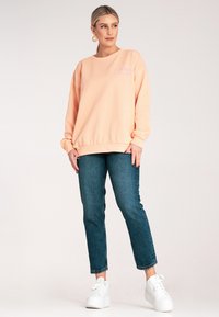 Brzoskwiniowa bluza z ribbowymi mankietami i dołem, z białym haftowanym napisem. Połączona z ciemnymi dżinsami i białymi trampkami na platformie.