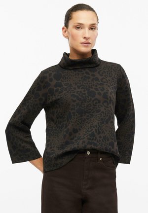 Frau trägt ein dunkelbraunes Langarmshirt mit Leopardenmuster und lockerem Rollkragen sowie dunkelbraune Hose vor weißem Hintergrund.