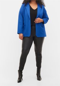 Blå blazer med slag över en svart topp, kombinerat med svarta läderbyxor och klackade ankelstövlar. Tyget verkar vara något texturerat.