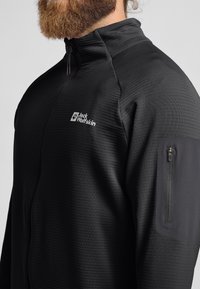Schwarze Zip-Jacke aus strukturiertem Stoff, mit einem Logo auf der Brust und einer mit Reißverschluss versehenen Tasche am Arm.