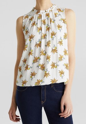 Blouse blanche sans manches à motif floral jaune et feuilles vertes, col froncé, portée avec un jean bleu foncé.