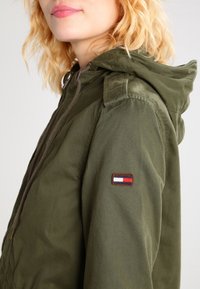 Giacca a vento verde oliva con cappuccio e chiusura zip, dotata di un piccolo logo ricamato sulla manica sinistra e un tessuto texture.