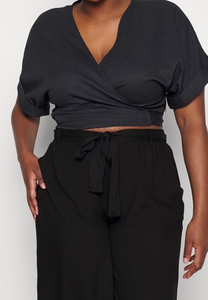 Kvinde iført en sort wrap-crop-top med korte ærmer og sorte højtaljede bukser med et bundet bælte i taljen.