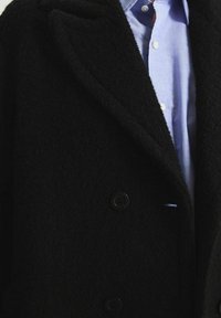 Manteau noir en laine avec une finition texturée, doté d'un design à double boutonnage, de deux boutons noirs et d'une chemise à col bleu clair en dessous.