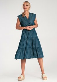 Sukienka z denimu w głębokim odcieniu teal z krótkimi rękawami, warstwowymi falbanami u dołu i wyraźnie zaznaczoną talią. Zestawiona z plecionymi sandałami na platformie.