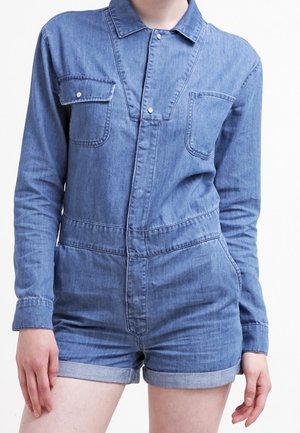 Salopette di denim in azzurro chiaro, con maniche lunghe, colletto, chiusura frontale con bottoni, due tasche sul petto e pantaloni corti arrotolati.