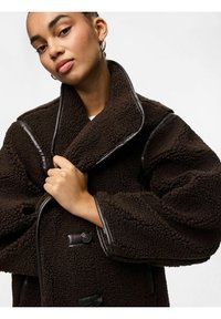 Rjav flisast plašč s puhasto teksturo, prevelikim ovratnikom in kontrastnimi usnjenimi robovi. Ima velike gumbe in sproščen kroj.