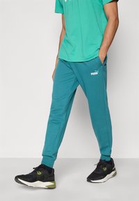 Τζόγκινγκ παντελόνι σε τυρκουάζ με ελαστικά τελειώματα, πλαϊνές τσέπες και λευκό λογότυπο Puma. Φορεμένο με πράσινο πουκάμισο και μαύρα αθλητικά παπούτσια.