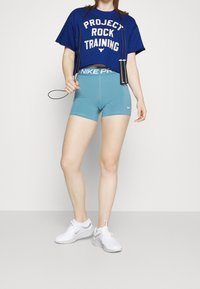 Blå kortärmad t-shirt med vit text, tillsammans med ljusblå Nike-shorts och vita sportskor. Hopprep hålls i höger hand.