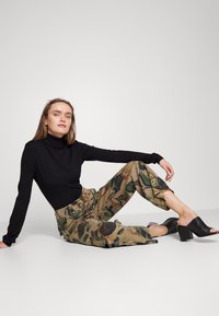 Suéter ajustado preto de gola alta, calças de cintura alta com padrão camuflado e mules pretas de salto aberto. Modelo sentado contra um fundo simples.