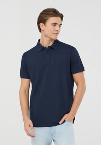 Granatowa koszulka polo z bawełny. Posiada klasyczny kołnierzyk, zapięcie na trzy guziki oraz krótkie rękawy. Brak wzorów lub akcentów.