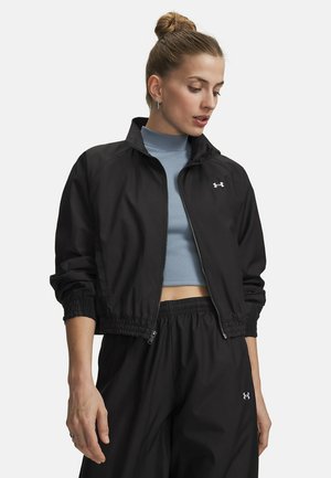 Frau mit Dutt trägt eine schwarze Zip-Up-Jacke über einem blauen, kurzen Rollkragenpullover und passende schwarze Hosen mit Logos auf Jacke und Hose.