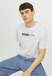Witte katoenen T-shirt met het "JACK & JONES" logo in het zwart, met een ronde hals en korte mouwen, gedragen met lichtblauwe denim jeans.