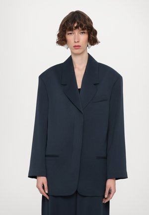 Granatowa, oversize żakiet wykonany z sztywnego materiału, z kołnierzem typu notch, pojedynczą kieszenią na piersi oraz dwiema przednimi kieszeniami o gładkiej fakturze.