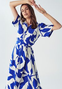 Žena v modro-bílé vzorované košilové šaty s krátkým rukávem, pózující s rukama vztyčenýma proti jednoduchému světlému pozadí.