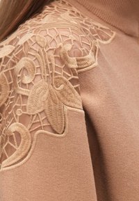 Bruine trui met een getextureerde overlay en ingewikkeld borduurwerk in een bloemenpatroon, met doorschijnende mesh accenten op de mouwen.