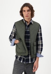 Tīkla struktūras zaļa veste ar augstu apkakli, ar rāvējslēdzēju un priekšējām kabatām, uzvilkta virs tumši zilā un krēmkrāsas šūnu krekliņa. Tekstūrēta auduma apdare.